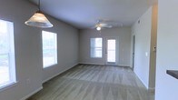 This single-floor space features an open living area finished with neutral-toned carpet flooring and light gray walls, creating a clean and versatile environment. A ceiling fan with an integrated light fixture is centrally positioned, ensuring efficient air circulation and ambient lighting throughout the room. Natural light enters generously through multiple windows fitted with horizontal blinds, as well as a glass-panel exterior door that opens to what appears to be an outdoor area such as a patio or balcony. The door and windows together create a bright, airy atmosphere while maintaining a connection to the outdoors. A decorative pendant light fixture is visible near the entrance, adding a subtle design element to the space. White baseboards provide a crisp contrast against the neutral carpeting and walls. The layout appears open and unobstructed, offering flexibility for various furniture arrangements. The overall ambiance is understated and functional, well-suited for everyday living. The combination of soft flooring, ample natural light, and simple finishes makes this a practical and comfortable space.
