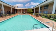 This single-story property showcases a classic brick exterior with notable architectural details, including prominent white colonial-style columns that frame the building's facade. The structure is arranged in a courtyard configuration, enclosing a generously sized rectangular swimming pool as the central focal point of the outdoor space.

The pool area is finished with traditional brick pavers that line the perimeter, providing a warm, cohesive aesthetic that complements the building's exterior. Several lounge chairs and seating arrangements are positioned poolside, offering residents a comfortable outdoor retreat. Mature trees and manicured shrubs are interspersed throughout the courtyard, adding greenery and shade to the environment.

The building features large multi-pane windows that allow natural light into the interior spaces while maintaining the architectural character of the facade. The surrounding urban backdrop, including a high-rise visible in the distance, suggests a central city location. Overall, the space conveys a well-maintained, community-oriented environment with a blend of classic architectural styling and practical outdoor amenities.
