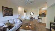 This single-floor residence offers an open-concept floor plan that seamlessly connects the living, dining, and kitchen areas, creating a spacious and cohesive environment ideal for everyday living and entertaining. The living room is anchored by a plush light-toned sofa, an ornate wooden coffee table, and a ceiling fan overhead, contributing to a comfortable and airy atmosphere. Rich hardwood flooring flows throughout the main living spaces, adding warmth and a sense of continuity. The dining area features a round table with elegant wrought-iron chairs, complemented by a decorative circular mirror and a hanging pendant light that adds a touch of sophistication. Natural light filters into the space through a window near the front entry, brightening the interior. The kitchen is visible through an open pass-through and is equipped with wooden cabinetry, modern appliances including a stainless steel microwave, and a raised breakfast bar with bar-height seating — a practical and inviting spot for casual dining. The front entryway is clean and functional, with a white paneled door that completes the welcoming feel of this thoughtfully designed home.