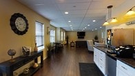This single-floor space is designed with an open-concept layout that seamlessly integrates multiple functional zones into one cohesive environment. The interior is anchored by rich, dark hardwood flooring that runs throughout, complemented by warm yellow-toned walls that create an inviting and comfortable ambiance.

The space features a generously sized lounge area furnished with a sofa and seating arrangement, centered around a wall-mounted flat-screen television — ideal for gatherings or relaxation. Adjacent to the lounge is a casual dining area equipped with bar-height tables and stools, providing a versatile spot for meals or informal meetings.

A kitchenette is positioned along one side of the space, outfitted with white cabinetry, dark granite countertops, and a sink — offering practical amenities in a clean, modern finish. Decorative elements such as tall vases, potted plants, and a console table add character and warmth to the entryway area.

Overhead, a drop-panel ceiling fitted with an abundance of recessed lighting ensures the entire space is evenly and brightly illuminated. Windows dressed with horizontal blinds allow for natural light control while maintaining privacy. The overall design strikes a balance between functionality and comfort, making this space well-suited for community use, hospitality, or professional settings.