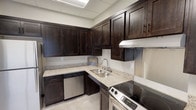 This single-floor space is centered around a fully equipped kitchen area that combines functionality with a refined aesthetic. The kitchen features rich, dark espresso-toned cabinetry with shaker-style doors that extend from floor to ceiling, providing generous storage throughout. Granite countertops in a light, speckled finish offer ample workspace and contrast elegantly against the dark cabinetry. Stainless steel appliances — including a full-size refrigerator, electric range with oven, built-in dishwasher, and microwave — complete the well-outfitted setup. A stainless steel backsplash behind the range adds a sleek, commercial-grade touch, while a double basin sink with a modern faucet is conveniently positioned along the counter. The flooring throughout is light-toned tile, lending a clean and cohesive look to the space. A suspended drop ceiling with recessed fluorescent panel lighting provides bright, even illumination. A large window fitted with horizontal blinds allows natural light to filter in and offers views of the surrounding neighborhood. An adjacent open area with neutral walls and additional cabinetry extends the functionality of the space, making it well-suited for use as a communal kitchen or employee break room.