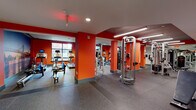This fitness center is designed to provide a comprehensive workout environment across a single floor, organized into distinct zones for cardio and strength training. The space is immediately distinguished by its bold orange-red accent walls and structural columns, which lend an energetic, motivating atmosphere throughout.

The cardio area features a row of treadmills positioned along large windows that admit natural light and offer views of the surrounding urban streetscape. Alongside the treadmills, users will find elliptical trainers, stationary bikes, and a rowing machine, all equipped with individual monitors. A dramatic full-wall photographic mural depicting a city bridge at dusk serves as a striking visual focal point in this section.

The strength training area is equipped with cable machines, a power rack, a flat bench, and a well-stocked dumbbell rack. Floor-to-ceiling mirrors line the walls, amplifying the sense of space and allowing users to monitor their form. Recessed lighting and linear LED fixtures provide consistent, even illumination throughout.

Durable speckled rubber flooring covers the entire facility, providing both safety and sound absorption. A separate adjoining room with hardwood flooring appears to offer additional activity space. The overall design balances functionality with a high-energy aesthetic.