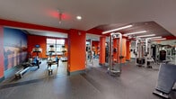 This fitness center is designed to provide a comprehensive workout environment across a single floor, organized into distinct zones for cardio and strength training. The space is immediately distinguished by its bold orange-red accent walls and structural columns, which lend an energetic, motivating atmosphere throughout.

The cardio area features a row of treadmills positioned along large windows that admit natural light and offer views of the surrounding urban streetscape. Alongside the treadmills, users will find elliptical trainers, stationary bikes, and a rowing machine, all equipped with individual monitors. A dramatic full-wall photographic mural depicting a city bridge at dusk serves as a striking visual focal point in this section.

The strength training area is equipped with cable machines, a power rack, a flat bench, and a well-stocked dumbbell rack. Floor-to-ceiling mirrors line the walls, amplifying the sense of space and allowing users to monitor their form. Recessed lighting and linear LED fixtures provide consistent, even illumination throughout.

Durable speckled rubber flooring covers the entire facility, providing both safety and sound absorption. A separate adjoining room with hardwood flooring appears to offer additional activity space. The overall design balances functionality with a high-energy aesthetic.
