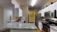 This single-floor residence offers a thoughtfully designed layout centered around a functional and stylish kitchen. The kitchen is a standout feature, showcasing granite countertops with a classic speckled finish that provide ample workspace for cooking and meal preparation. White shaker-style cabinetry lines the walls, offering generous storage while contributing to a bright, clean aesthetic. The backsplash features neutral tile work that complements the overall color palette of warm greige tones throughout the space. Stainless steel appliances, including a refrigerator and a gas range, add a modern touch and enhance the kitchen's practicality. A deep sink is conveniently positioned along the countertop, making the space as functional as it is visually appealing. Light hardwood flooring runs throughout, adding warmth and continuity to the interior. The layout flows naturally toward an adjoining room, visible through an interior doorway that hints at a connected and cohesive floor plan. Overhead lighting ensures the kitchen remains well-lit, creating an inviting atmosphere suitable for both everyday living and entertaining.