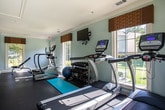 This single-story fitness center in Peachtree Corners, Georgia, offers a well-equipped exercise space designed to support a range of workout routines. The interior is finished with soft blue-green walls, white crown molding, and dark rubber flooring throughout, creating a clean and functional environment.

The main workout area is generously proportioned, accommodating multiple cardio machines including True-brand treadmills and an elliptical trainer. A rack of dumbbells, a stability ball, a yoga mat, and an adjustable weight bench provide options for strength and flexibility training. A wall-mounted flat-screen television adds convenience for entertainment during workouts.

Natural light fills the space through several large windows adorned with warm, patterned valances. A standout window overlooks an outdoor pool area, offering a pleasant view during cardio sessions. An exterior door with a glass panel provides additional light and direct outdoor access.

A small entry corridor connects the fitness room to an adjacent space and includes a water cooler station for hydration convenience. The overall layout is practical and accessible, with an exit-marked door ensuring safe egress. Crown molding and consistent finishes throughout lend a polished, well-maintained character to the facility.