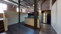 This industrial-style loft showcases the character of a thoughtfully converted historic building, blending raw architectural elements with modern finishes throughout. The open-plan living and kitchen area is anchored by striking exposed brick walls, concrete ceilings with visible steel ductwork, and bold structural columns that speak to the building's industrial heritage. Dark hardwood floors run through the main living space, complementing the rugged aesthetic.

The kitchen is a standout feature, offering dark cabinetry, granite countertops, a central island with additional counter seating, and stainless steel appliances including a refrigerator, range, and microwave. Vintage-style pendant lights hang above, enhancing the loft's industrial charm. A staircase leads to an upper landing area, adding a sense of vertical dimension to the layout.

The bedroom features original exposed brick walls and large windows that invite generous natural light, with plush carpeting providing a warm contrast to the raw surroundings. Two full bathrooms are finished in clean, neutral tones with tub/shower combinations and tile flooring. Together, these elements create a space that is both historically rich and comfortably livable.