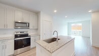 This well-appointed residence at The Enzo at Ariston showcases a thoughtfully designed single-floor layout with a clean, contemporary aesthetic. The open-concept kitchen is a standout feature, centered around a large island with granite countertops and an integrated sink. White shaker-style cabinetry extends to the ceiling, providing ample storage, while a full suite of stainless steel appliances — including a refrigerator with ice maker, electric range, over-the-range microwave, and dishwasher — completes the space. Light hardwood-style flooring runs throughout the main living areas, enhancing the bright, airy feel created by recessed lighting and natural light from large windows. The open living area flows seamlessly toward a private balcony, offering an inviting outdoor connection. Bedrooms are carpeted for comfort, with one featuring a ceiling fan and a window with exterior views. The bathroom includes a granite-topped vanity, a combination tub and shower, and direct access to a walk-in closet with built-in shelving. Throughout the unit, crisp white walls, clean lines, and modern fixtures come together to create a fresh, move-in-ready living environment.