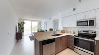 This single-floor residence showcases a clean, contemporary design that maximizes both style and functionality. The open-concept layout seamlessly connects the kitchen and living area, creating a bright and airy atmosphere throughout. The kitchen is well-appointed with two-tone cabinetry — combining crisp white upper cabinets with warm wood-finish lower cabinets — complemented by light quartz countertops and a modern undermount sink. Stainless steel appliances, including a dishwasher and range, add a polished, professional touch. A peninsula counter extends the workspace while also serving as a casual dining or gathering spot, enhancing the open flow of the space. Light-toned flooring runs throughout, reflecting natural light and contributing to the home's spacious feel. Large floor-to-ceiling sliding glass doors lead to an outdoor balcony, where lush tropical greenery and open water views can be enjoyed. The living area benefits greatly from this indoor-outdoor connection, bathed in natural light. The overall ambiance is modern and refined, with a neutral color palette and thoughtful design details that create a welcoming and comfortable environment.