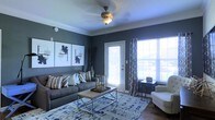 This single-story space features a thoughtfully designed living area that combines modern aesthetics with comfortable functionality. The living room is immediately distinguished by its deep slate-blue accent walls, which create a bold yet sophisticated atmosphere. Crown molding adds an elegant architectural detail, framing the space with a refined finish, while a textured ceiling and a ceiling fan with a classic light fixture provide both practical comfort and visual interest.

Natural light floods the room through large windows dressed with patterned curtains, as well as a glass-paneled door that leads to an outdoor area. The bright, airy quality of the light contrasts beautifully with the rich wall color, giving the space a dynamic and inviting character. Hardwood-style flooring anchors the room, complemented by a large area rug in blue and grey tones that defines the seating arrangement.

The furnishings include a plush grey sofa adorned with layered accent pillows, a modern wood-and-metal coffee table, and a light-toned armchair. A gallery wall of abstract artwork adds a curated, artistic touch, completing the polished, contemporary feel of the space.
