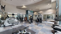 This modern fitness center is thoughtfully designed across a single floor, offering two distinct functional zones for a comprehensive workout experience. The main exercise area boasts soaring ceilings with a curved architectural detail overhead, recessed lighting, and large windows that flood the space with abundant natural light and views of the surrounding outdoors. The floor is finished with durable, interlocking tile in a slate-toned pattern, providing both durability and visual appeal. This primary zone is extensively equipped with a wide array of strength-training machines, cable systems, multi-functional weight stations, adjustable benches, and a full rack of dumbbells, all complemented by wall-to-wall mirrors and mounted flat-screen televisions. A separate studio room features warm hardwood flooring, full-length mirrors along one wall, ceiling fans, and large windows that bring in natural light — ideal for cycling, stretching, or group fitness activities. Spinning bikes are positioned along the perimeter, making the most of the open floor space. The overall ambiance of the facility is clean, bright, and contemporary, with a neutral color palette of grays and whites accented by a teal feature wall, creating a motivating and professional environment suited for a variety of fitness goals.