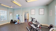 Located at 500 Carlen Ave in Lexington, South Carolina, this fitness center spans a single floor divided into two interconnected exercise rooms, offering a comprehensive workout environment for residents. The space is finished with durable rubber flooring throughout and painted in a calming light aqua hue, creating an energizing yet relaxed atmosphere conducive to exercise.

The first exercise room is dedicated primarily to cardio training and functional fitness. It houses a row of cycling bikes alongside balance and stability tools, including medicine balls, a ball rack, and various weighted accessories. Motivational framed artwork — featuring the words "inhale" and "exhale" — adds a mindful touch to the walls. Natural light filters in through shuttered windows, complemented by overhead panel lighting and a ceiling fan.

The second exercise room expands the facility's offerings with a broader range of equipment. Elliptical trainers, a treadmill, and a cable resistance machine provide options for both cardiovascular and strength training. A dumbbell rack with a weight bench, wall-mounted mirrors, and a ceiling-mounted television round out the amenities. Large windows along one wall allow ample natural light, and a direct exterior door provides convenient access. Together, the two rooms form a fully functional fitness facility suited for a variety of training routines.