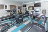 Located at 6033 Fountain Pointe in Grand Blanc, Michigan, this well-appointed fitness center offers a thoughtfully designed, single-level layout divided into two distinct functional zones. The cardio and machine area features bold, geometric-patterned carpet tiles in teal, gray, and white, providing a dynamic visual foundation for an array of equipment including treadmills, stationary bikes, an elliptical, and a recumbent cycle. A striking dark stacked-stone accent wall serves as the focal point of this zone, lending an upscale, modern aesthetic, while recessed lighting illuminates the space evenly throughout. Multiple wall-mounted flat-screen televisions are positioned for viewing during workouts, and a built-in water fountain station with a bottle-filling option adds practical convenience. The adjacent functional training area transitions to smooth hardwood-style flooring and features a wall-mounted television, medicine ball rack, stability balls, and balance training tools, offering ample open space for stretching and functional movement exercises. Recessed ceiling lights maintain consistent brightness across both areas. Interior French doors connect the two zones seamlessly, and the overall neutral gray palette with contrasting accent elements creates a clean, motivating environment well-suited for a comprehensive fitness routine.