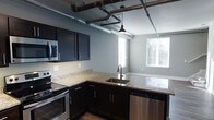 This two-floor space blends industrial character with modern finishes to create a distinctive and functional living environment. The kitchen is a standout feature, showcasing dark espresso cabinetry paired with light granite countertops that provide a striking contrast. Stainless steel appliances, including a range, built-in microwave, and dishwasher, add a contemporary touch, while a deep undermount sink with a sleek gooseneck faucet anchors the L-shaped counter layout. Overhead, exposed ductwork and industrial-style pipes run across the ceiling, adding authentic loft character that is complemented by a hanging pendant light with a vintage-inspired shade. The cool grey walls and light-toned flooring keep the space feeling open and airy, while multiple windows allow generous natural light to pour in from multiple angles. The open sightline from the kitchen into the adjacent living or dining area enhances the sense of spaciousness throughout the main level. Spanning two floors, the layout offers a layered living experience that combines the raw, urban appeal of industrial design with the comfort and polish of modern finishes — making it a compelling and well-appointed space.