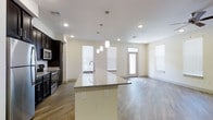 This single-floor space showcases a thoughtfully designed open-concept layout that seamlessly connects the kitchen and living area. The kitchen is a standout feature, anchored by a large center island topped with granite countertops and equipped with built-in electrical outlets for added convenience. Pendant lighting hangs above the island, providing focused illumination while adding a contemporary design element. Dark cabinetry contrasts cleanly with the light-toned walls and granite surfaces, and stainless steel appliances, including a microwave and range, complete the functional cooking space.

Wood-style flooring extends throughout the main living area, adding warmth and continuity to the open floor plan. Recessed lighting in the ceiling supplements the generous natural light streaming in through multiple windows dressed with horizontal blinds. The living area is generously proportioned, offering flexible arrangement possibilities. A entry door with an adjacent window further brightens the space. The overall ambiance is clean, modern, and welcoming, with a neutral color palette that enhances the sense of openness throughout.