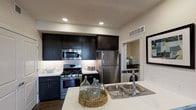 This thoughtfully designed single-floor residence offers a seamless blend of modern aesthetics and practical functionality. The open-concept living and dining area is anchored by rich, wide-plank hardwood-style flooring that extends throughout, complemented by neutral-toned walls that create a bright and welcoming atmosphere. A ceiling fan provides comfort overhead, while large sliding glass doors flood the space with natural light and open onto a private balcony, offering views of the surrounding area. A full-length mirror with a bold black frame adds a striking decorative touch to the living space.

The kitchen is a standout feature, showcasing sleek dark cabinetry that reaches to the ceiling, paired with crisp white countertops and a suite of stainless steel appliances including a gas range, built-in microwave, and refrigerator. Recessed lighting illuminates the space, while a double-basin sink at the island provides additional prep space. Conveniently accessible from the kitchen, an in-unit laundry area with a washer is neatly tucked behind a sliding door, adding everyday practicality. Together, these elements create a cohesive, contemporary living environment suited for modern lifestyles.