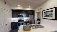 This thoughtfully designed single-floor residence offers a seamless blend of modern aesthetics and practical functionality. The open-concept living and dining area is anchored by rich, wide-plank hardwood-style flooring that extends throughout, complemented by neutral-toned walls that create a bright and welcoming atmosphere. A ceiling fan provides comfort overhead, while large sliding glass doors flood the space with natural light and open onto a private balcony, offering views of the surrounding area. A full-length mirror with a bold black frame adds a striking decorative touch to the living space.

The kitchen is a standout feature, showcasing sleek dark cabinetry that reaches to the ceiling, paired with crisp white countertops and a suite of stainless steel appliances including a gas range, built-in microwave, and refrigerator. Recessed lighting illuminates the space, while a double-basin sink at the island provides additional prep space. Conveniently accessible from the kitchen, an in-unit laundry area with a washer is neatly tucked behind a sliding door, adding everyday practicality. Together, these elements create a cohesive, contemporary living environment suited for modern lifestyles.