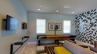 This single-floor space is designed as a social and recreational lounge, offering a welcoming environment for leisure and entertainment. The room is anchored by a full-length wooden shuffleboard table, serving as a central focal point for group activities and casual play. A wall-mounted flat-screen TV is positioned on one side, providing additional entertainment options for guests.

The interior features a light, neutral color palette with soft blue-gray walls that create a relaxed and inviting atmosphere. Recessed lighting in the white ceiling ensures the space is well-lit throughout. Multiple windows fitted with horizontal blinds allow natural light to filter in, brightening the room and giving it an open, airy feel.

Seating includes a patterned sofa and accent ottoman in bold yellow, adding a vibrant pop of color and modern flair to the space. A striped area rug grounds the seating arrangement and adds visual texture to the hardwood-style flooring. Framed abstract artwork adorns the walls, complementing the contemporary aesthetic.

Overall, this lounge space strikes a balance between functionality and style, making it well-suited for social interaction, recreation, and relaxed gatherings in a thoughtfully designed, single-level setting.