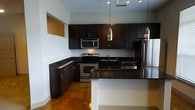 This single-floor space offers a well-organized layout that balances open communal areas with more defined private rooms. The kitchen serves as a standout feature, showcasing dark espresso cabinetry that extends to the upper walls, providing generous storage throughout. Dark granite countertops complement the cabinetry and contrast with the white walls, while stainless steel appliances — including a range, built-in microwave, dishwasher, and refrigerator — contribute a sleek, modern aesthetic. A breakfast bar peninsula defines the kitchen's boundary and creates a natural transition into the adjoining living area. Elegant pendant lights hang above the peninsula, while a track lighting system illuminates the kitchen workspace. The flooring throughout the main living and kitchen areas consists of polished, warm-toned stained concrete, lending an industrial yet inviting character to the space. The open living area benefits from natural light provided by a window along one wall. Additional rooms are accessible toward the rear of the layout, including what appears to be a bathroom and a separate enclosed room, offering functional separation within the overall floor plan. The white walls and high ceilings enhance the sense of openness throughout.