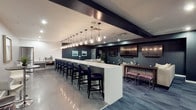 The Quincy Media Room is a thoughtfully designed single-floor space that blends modern aesthetics with functional comfort. The centerpiece of the room is an expansive white bar counter stretching the length of the space, lined with a row of dark upholstered bar stools that invite guests to settle in. Overhead, a striking series of globe pendant lights hang in a continuous line above the bar, casting warm, ambient light across the room and serving as a defining design feature. Dark accent beams cross the ceiling, adding architectural contrast against the lighter ceiling panels. The walls are finished in deep, sophisticated tones, complemented by framed artwork that adds a refined touch. Multiple flat-screen televisions are mounted throughout the room, ensuring clear sightlines from virtually every seat — making this space well-suited for media viewing and entertainment. On the opposite side of the bar, a set of plush pink ottomans arranged around low tables offer an alternative, relaxed seating option. The polished tile flooring reflects the room's lighting, enhancing the sleek, contemporary atmosphere. Overall, the Quincy Media Room delivers a polished and versatile environment designed for both casual socializing and organized media events.