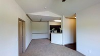 This single-story residence showcases a thoughtfully designed open-concept layout that connects the living area and kitchen in a cohesive, functional flow. The living room draws the eye upward with its vaulted, angled ceiling, which lends a sense of spaciousness and architectural character to the space. Crown molding trim accents the transition between walls and ceiling, adding a refined finishing touch. Neutral-toned carpet covers the living area floor, contributing to a warm and inviting atmosphere.

A half-wall peninsula separates the kitchen from the living space while maintaining visual openness throughout. The kitchen features dark cabinetry, black appliances, and under-cabinet lighting that provides functional illumination for the cooking area. The counter space along the peninsula offers casual seating or serving potential.

A doorway visible to the right leads to an adjacent room with hardwood-style flooring, suggesting a distinct functional zone such as a dining area or additional living space. An entry door with a mirrored closet is visible on the left side of the living room. Overall, the space balances practicality with clean, straightforward design elements suited to everyday living.