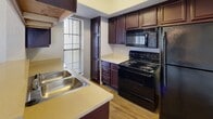 This single-floor residence offers a practical and comfortable layout designed for everyday living. The living room serves as the heart of the home, centered around a classic brick fireplace flanked by built-in shelving units — an ideal spot for display or storage. Soft carpet underfoot and a ceiling fan overhead add to the room's comfort, while a large window allows natural light to fill the space. An adjacent area, also carpeted and featuring a floor-to-ceiling mirror wall, provides additional flexible living space with direct access to the outdoors through a glass-paned door. The kitchen is well-appointed with rich dark wood cabinetry offering generous storage, light-toned countertops, a double-basin stainless steel sink, and a full suite of black appliances including an electric range, over-the-range microwave, and refrigerator. Light hardwood-style flooring grounds the kitchen's warm aesthetic. The bathroom is thoughtfully designed with white cabinetry, a single-basin vanity with a light countertop, a large mirror with overhead vanity lighting, and a full tub and shower combo finished with neutral tile. The textured ceilings throughout lend the space a classic character consistent across all rooms.
