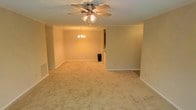 This single-floor space presents an open and functional layout anchored by a generously sized living area with wall-to-wall carpeting in a warm beige tone, contributing to an inviting and comfortable atmosphere. The main living space is illuminated by a ceiling fan with an integrated light fixture, providing both air circulation and ambient lighting. Crown molding lines the upper perimeter of the walls, adding a subtle architectural detail that enhances the overall finish of the space.

Toward the back of the living area, a decorative chandelier marks a transition into a secondary zone, which opens into what appears to be a dining or additional living area, suggesting a flowing, connected layout ideal for everyday living and entertaining. The walls are painted in a soft, neutral tone that complements the carpeting and contributes to the warm ambiance throughout.

A partial wall and pass-through opening hint at a kitchen area beyond, indicating an accessible and practical design. White baseboards provide clean contrast against the neutral walls and flooring. The space is well-proportioned, with a sense of openness that makes it feel roomy and versatile for a variety of furniture arrangements and uses.