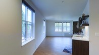 This single-floor space is characterized by its open-concept layout and an abundance of natural light. Multiple large windows with roller shades line the walls, offering views of surrounding greenery and allowing daylight to flood the interior throughout the day. The clean white walls and smooth ceilings create a bright, airy atmosphere that enhances the sense of openness.

The flooring throughout is a light-toned hardwood-style material that adds warmth while complementing the neutral color palette. The main living area is generously proportioned, providing flexible space for a variety of furniture arrangements. A wall-mounted HVAC unit ensures climate control convenience.

Toward one end of the space, a kitchen area is partially visible, featuring dark wood cabinetry paired with a light countertop surface — a contrast that adds visual interest to the otherwise minimalist design. A small area rug near the kitchen zone adds a subtle decorative touch.

Overall, the space maintains a clean, modern aesthetic with practical features, making it a well-lit and functional environment suited for comfortable everyday living.