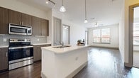 This contemporary single-floor residence in Katy, Texas, offers a thoughtfully designed open-concept layout with modern finishes throughout. The kitchen serves as the heart of the home, anchored by a large center island with a crisp white quartz countertop, an undermount sink, and pendant lighting overhead. Dark walnut-toned cabinetry pairs elegantly with a mosaic tile backsplash, and a full suite of stainless steel appliances—including a French door refrigerator, dishwasher, microwave, and range—completes the space. Rich, dark hardwood-style flooring flows seamlessly from the kitchen into the open living area, which is brightened by multiple windows and equipped with a ceiling fan. Additional living spaces feature light-toned wood-style flooring and generous window coverage, flooding each room with natural light. The bedrooms are carpeted for comfort, with ceiling fans and large windows offering views of the surrounding neighborhood. A spacious walk-in closet with built-in shelving and dual hanging rods provides ample storage. The bathroom is finished with a wood-grain vanity, quartz countertop, large framed mirror, and a tiled tub-shower combination. The overall ambiance is clean, modern, and move-in ready.