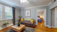 This single-floor space offers a thoughtfully designed layout connecting key living areas through a cohesive aesthetic of soft gray walls, warm hardwood flooring, and classic crown molding throughout. The living room serves as a welcoming central space, featuring a ceiling fan with light fixture, large windows dressed in charcoal drapes that admit generous natural light, and ample room for comfortable seating arrangements around a solid wood coffee table. Framed artwork accents the walls, adding a cultivated touch to the relaxed atmosphere.

The bedroom is well-proportioned, accommodating a queen-sized bed with a decorative latticed headboard. Natural light filters through windows fitted with blinds and curtain panels, complemented by warm-toned hardwood floors and a ceiling fan overhead. Rustic wooden nightstands complete the room's grounded, restful feel.

The kitchen is efficiently arranged with white cabinetry, dark countertops, white subway tile backsplash, a double-basin sink, and a patterned tile floor. Natural light from a window brightens the workspace. The unit includes two bathrooms: one featuring a corner glass-enclosed shower stall, pedestal sink, and oval mirror; the other a full bath with a shower-tub combination, toilet, and white tile flooring.