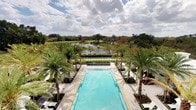 This expansive outdoor amenity space showcases a generously sized rectangular swimming pool as its centerpiece, finished in a luminous aqua tone that reflects the open Florida sky above. The pool deck is clad in light-toned stone pavers, lending a clean, sophisticated aesthetic throughout. Flanking both sides of the pool are rows of mature, towering palm trees that provide a tropical canopy and a sense of privacy, while lounge chairs are neatly arranged along the perimeter for relaxation. A water feature element is positioned at the far end of the pool, adding a subtle dynamic accent to the overall design. Beyond the pool, meticulously trimmed hedgerows transition into a serene pond, its calm surface mirroring the surrounding greenery. Further in the distance, a lush golf course stretches across the landscape, offering panoramic views of rolling fairways and mature tree lines. The combination of the resort-caliber pool, curated tropical landscaping, and unobstructed natural vistas creates an ambiance that is both refined and tranquil — ideal for leisure and outdoor enjoyment.