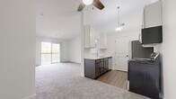 This single-floor, 2-bedroom, 1-bathroom residence offers a clean and modern layout designed for comfortable everyday living. The open-concept kitchen and living room serve as the heart of the home, with the kitchen showcasing white upper cabinetry, dark lower cabinets, stainless steel appliances including a refrigerator, range, and over-the-range microwave, and wood-look vinyl flooring. A pendant light fixture illuminates the space, which connects seamlessly to the carpeted living area via a pass-through window. The living room is generously sized, featuring a ceiling fan, neutral carpet, and sliding glass door access to a private balcony with outdoor views. Both bedrooms are carpeted and bathed in natural light through sliding windows that frame views of lush greenery. The primary bedroom includes a walk-in closet and convenient access to the full bathroom. The full bathroom features wood-look vinyl flooring, a dark vanity with a white countertop, and a tub-shower combination. A hallway connects the private quarters to the main living spaces, and the textured ceilings and bright white walls throughout contribute to the home's clean, cohesive aesthetic.