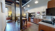 Located at 900 Goss Ave in Louisville, KY, this 2-bedroom, 2-bathroom loft-style unit masterfully blends industrial heritage with modern comfort. The open living and kitchen area is defined by soaring ceilings with exposed ductwork, original hardwood floors, and dramatic floor-to-ceiling arched windows that flood the space with natural light. The exposed brick walls throughout add rich texture and character, speaking to the building's storied past.

The kitchen features warm wood cabinetry, granite countertops, stainless steel appliances including a full-size refrigerator and dishwasher, and open metal shelving for a clean, functional aesthetic. A rustic wood dining table with bar seating anchors the combined kitchen and dining area.

The primary bedroom retains the industrial charm with exposed brick walls, original windows, and rich hardwood floors, offering a spacious and comfortable retreat. The second bedroom showcases a distinctive green barn-style sliding door leading to a closet, adding a playful design element. Both bathrooms are well-appointed, with one featuring a tiled tub-shower combination. Throughout the unit, the interplay of raw materials and contemporary finishes creates a cohesive, urban-loft ambiance unique to Louisville's architectural character.