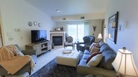 This single-floor residence offers a thoughtfully designed and warmly decorated living space with a distinct eclectic charm. The main living area is the focal point of the home, centered around a classic fireplace with a light wood mantel that serves as a natural gathering point. A large sliding glass door floods the room with natural light, creating a bright and inviting atmosphere while offering a view to the outdoors.

The living room is generously proportioned, accommodating a plush sofa adorned with layered throw pillows in warm tones, a tufted ottoman serving as a central coffee table, and a pair of accent chairs — one upholstered in a bold animal print and another in rich dark leather — that add visual interest and personality to the space. A large flat-screen television is mounted on a rustic media console, balancing entertainment functionality with stylish design.

Decorative wall accents, including a sunburst mirror arrangement and a statement wall sconce, contribute to the room's curated aesthetic. Soft carpeting and a dark area rug add texture and comfort underfoot. The overall ambiance is cozy yet stylish, blending traditional furnishings with eclectic accents to create a lived-in, welcoming environment ideal for both relaxing and entertaining.
