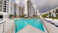 This contemporary residential complex showcases a striking modern architectural design, characterized by clean white facades, bold geometric lines, and rows of private balconies that rise across multiple stories. The standout feature of the property is its expansive resort-style swimming pool, which serves as the centerpiece of the outdoor communal area. The pool's vivid turquoise waters are complemented by light stone tile decking, offering ample space for lounging with poolside chairs and chaise lounges positioned for relaxation. Lush tropical palm trees and manicured greenery are thoughtfully integrated throughout the courtyard, evoking a warm, South Florida ambiance. The multi-wing building layout wraps around the pool area, creating an intimate yet open-air atmosphere. Each residential floor features generously sized balconies, providing elevated views of the pool and surrounding landscape. The architectural design emphasizes a seamless indoor-outdoor lifestyle, with the communal outdoor space designed to encourage social gathering and leisure. The overall aesthetic is modern and polished, reflecting a resort-inspired living experience within an upscale residential setting.