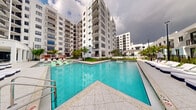 This contemporary residential complex showcases a striking modern architectural design, characterized by clean white facades, bold geometric lines, and rows of private balconies that rise across multiple stories. The standout feature of the property is its expansive resort-style swimming pool, which serves as the centerpiece of the outdoor communal area. The pool's vivid turquoise waters are complemented by light stone tile decking, offering ample space for lounging with poolside chairs and chaise lounges positioned for relaxation. Lush tropical palm trees and manicured greenery are thoughtfully integrated throughout the courtyard, evoking a warm, South Florida ambiance. The multi-wing building layout wraps around the pool area, creating an intimate yet open-air atmosphere. Each residential floor features generously sized balconies, providing elevated views of the pool and surrounding landscape. The architectural design emphasizes a seamless indoor-outdoor lifestyle, with the communal outdoor space designed to encourage social gathering and leisure. The overall aesthetic is modern and polished, reflecting a resort-inspired living experience within an upscale residential setting.