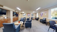 Located at 300 E Marshall St in West Chester, PA, this single-floor community lounge is a spirited gathering space designed with an unmistakable Philadelphia sports theme. The walls are adorned with prominent logos and graphics representing the Eagles, 76ers, Phillies, Flyers, and Philadelphia Union, creating an engaging and lively atmosphere throughout the room.

The open-plan layout offers generous, well-organized seating arrangements featuring navy blue upholstered armchairs grouped around light wood circular and rectangular tables, providing ample space for socializing and relaxation. The patterned, multi-tonal carpeting adds visual texture and warmth underfoot, complementing the neutral beige walls and white crown molding that frame the room.

Recessed and flush-mounted fluorescent ceiling lights ensure bright, even illumination throughout. On one wall, a built-in wood-cabinet kitchenette is equipped with counter space, a refrigerator, and a wall-mounted flat-screen television, making the space practical for gatherings and events. A second wall-mounted TV is also present, enhancing the room's entertainment capabilities.

A glass-partitioned entryway with automatic sliding doors connects the lounge to the exterior, allowing natural light to filter in and providing clear sightlines to the surrounding greenery. A dedicated alcove area near the entrance offers additional intimate seating, decorated with team logos and windows overlooking the outdoors.