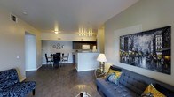 This single-floor residence is designed around an open-concept floor plan that integrates the living, dining, and kitchen areas into one cohesive, flowing space. The living area is anchored by a tufted gray sofa accented with decorative pillows in navy and yellow, complemented by a sleek glass-top coffee table and a stylish table lamp that adds warm ambient lighting. A large framed cityscape artwork adorns the wall, serving as a bold focal point. The kitchen features dark cabinetry contrasted by a white microwave and appliances, with a raised breakfast bar that separates it from the rest of the open space while maintaining visual connectivity. Track lighting above the kitchen illuminates the workspace effectively. Adjacent to the kitchen, the dining area accommodates a round table with modern chairs, decorated with a set of circular wall accents. Dark hardwood-style flooring runs throughout, adding warmth and continuity to the entire space. The neutral-toned walls and recessed ceiling lights contribute to a clean, contemporary ambiance that feels both functional and inviting.
