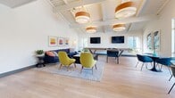 Located in Tustin, California, this single-floor community lounge space offers a thoughtfully designed environment ideal for gathering and recreation. The living area is the heart of the space, characterized by dramatic vaulted ceilings with exposed white-painted beams and warm wood accents. Large pendant light fixtures with wood-trimmed shades cast a welcoming glow throughout. Light wood-plank flooring runs seamlessly across the entire space, lending a clean and cohesive aesthetic. Seating areas are arranged around patterned area rugs, with vibrant yellow accent chairs and a navy sectional sofa creating a lively, social atmosphere. Wall-mounted flat-screen televisions, a foosball table, and a shuffleboard table add dynamic recreational elements. Expansive windows along the perimeter flood the space with natural light and provide views of the surrounding area. The kitchen is outfitted with sleek grey cabinetry, white quartz countertops, a white subway tile backsplash, a stainless steel range hood over a built-in cooktop, an under-counter microwave, and a separate wet bar area featuring a stainless steel sink and beverage refrigerator — all designed for functional, communal use.