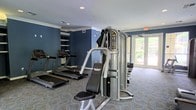 This fitness center offers a practical and well-organized space designed to support a full workout routine. The room features deep navy blue accent walls paired with crisp white crown molding, creating a clean and focused atmosphere. Recessed lighting in the ceiling provides consistent illumination throughout the space, complemented by large glass doors along one wall that invite natural light and offer views of the greenery outside.

The equipment layout is thoughtfully arranged to maximize floor space. Cardio equipment, including multiple treadmills, lines one side of the room, while a multi-station strength training machine occupies a central position, offering a variety of resistance exercises. Open shelving built into the wall provides convenient storage for personal items or accessories. The floor is covered in a patterned, low-pile carpet that adds both comfort and durability underfoot.

Crown molding details and the well-maintained finishes throughout give the space a polished appearance that elevates it beyond a standard gym setup. The natural light streaming through the glass doors enhances the overall ambiance, making the environment feel open and inviting. This fitness center is well-suited for residents or guests looking for a convenient and comfortable place to stay active.