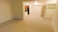 This single-floor space features an open layout that connects the main living area to the kitchen, creating a cohesive and functional flow throughout. The living area is finished with neutral-toned wall-to-wall carpeting, lending a clean and understated look to the space. Soft, light-colored walls throughout contribute to a bright and airy ambiance, enhanced by ceiling lighting that evenly illuminates the room.

The kitchen is accessible through an open pass-through or partial wall, offering visibility between the two spaces. It features warm wood cabinetry, dark countertops, and black appliances, including a full-size refrigerator. The kitchen area transitions to a non-carpeted floor, distinguishing it functionally from the living space.

Adjacent to the main living area, a doorway leads to an additional room, suggesting further private or functional spaces beyond what is immediately visible. The open design and connected rooms make the space feel practical and versatile, suitable for a variety of furniture arrangements and everyday living needs.