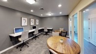 The business center at Lake Castleton is a thoughtfully designed, single-floor workspace that balances functionality with modern aesthetics. The space is finished in a contemporary gray palette, with textured ceilings, recessed lighting, and flush light fixtures that provide consistent, well-distributed illumination throughout.

The main working area features multiple white marble-topped desks arranged along the walls, each equipped with computers and ergonomic chairs, offering ample room for individual work. Open shelving with black metal brackets lines one wall alongside gray lower cabinetry with a marble-style countertop, providing storage and a dedicated area for office equipment such as a printer.

Natural wood round tables paired with upholstered chairs are positioned throughout the space, creating casual seating for collaborative discussions or informal meetings. Framed botanical artwork adorns the walls, adding a refined decorative touch.

Frosted glass panels and a glass door separate a private room within the suite, maintaining an open visual connection while ensuring acoustic privacy. Plush patterned carpet flooring ties the space together, contributing to a quiet, professional atmosphere well-suited for focused work or resident use.