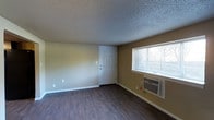 This single-floor space presents a straightforward, functional layout centered around an open living area. The room features durable, dark hardwood-style flooring that contrasts with the warm beige-toned walls, creating a simple yet cohesive aesthetic. A textured popcorn ceiling adds a classic touch overhead.

Natural light enters through a horizontal window fitted with horizontal blinds, offering a view of the outdoors. Beneath the window sits a wall-mounted air conditioning unit, providing climate control directly within the living space. A standard white panel door serves as the main entry point, positioned along one wall and complemented by clean white baseboards that frame the room neatly.

Electrical outlets are distributed along the walls, ensuring practical utility throughout the space. The room is unfurnished, allowing a clear view of its full dimensions and architectural simplicity. Overall, the space delivers a no-frills, clean environment well-suited for functional, everyday living.