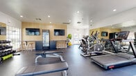 This fitness center is a thoughtfully designed, single-floor space that balances functionality with a clean, modern aesthetic. The interior features recessed lighting throughout, smooth white ceilings, and durable dark flooring laid with interlocking gym mats in key workout zones, providing both safety and comfort underfoot.

The strength training area is well-appointed with a multi-functional cable machine, a dumbbell rack stocked with a full range of free weights, and multiple flat and adjustable weight benches. Full-length mirrors span an entire wall, enhancing the sense of space and allowing users to monitor their form during exercise.

The cardio section houses an array of machines, including ellipticals, stair climbers, and treadmills, positioned to take advantage of natural light from large exterior-facing windows and glass doors that offer views of the surrounding area.

A standout feature is the dedicated "Hydration Station," set against a warm wood-panel accent wall. This area includes a glass-door beverage refrigerator, built-in navy cabinetry, a countertop workspace, and natural wood bar stools beneath wall-mounted flat-screen TVs. Stability balls and resistance accessories are also available. The space is bright, open, and designed to support a comprehensive fitness routine.