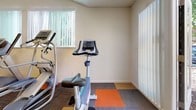 Located at 6430 Verner Ave in Sacramento, California, this single-floor fitness center offers a well-equipped workout space designed to support a range of exercise routines. The room features a distinctive multi-toned carpet tile flooring in a bold patchwork pattern of orange, gold, and gray — adding a vibrant, energetic feel to the space.

The facility is outfitted with a solid selection of exercise equipment, including a Precor elliptical machine, a treadmill, an upright stationary bike, and a multi-function cable weight station paired with an adjustable workout bench. This combination supports both cardiovascular and strength training needs.

The space benefits from natural light filtering through several windows dressed with vertical blinds, contributing to an open and airy atmosphere. A section of the room features vaulted ceilings with a skylight panel, enhancing the sense of volume. Recessed lighting and a flush panel light provide additional illumination throughout.

Climate comfort is maintained by a wall-mounted ductless mini-split HVAC unit, ensuring year-round temperature control. A water cooler is also present near the entrance, adding a practical convenience for users. The neutral warm-toned walls keep the environment clean and focused, making this a functional and comfortable space for daily fitness activity.
