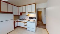 Located at 2055 Castle Ave N in North St. Paul, Minnesota, this single-floor unit offers a straightforward and functional layout across several well-defined rooms. The floor plan, visible in the 3D model, reveals a practical arrangement of living, sleeping, kitchen, and bathroom spaces connected by a clear interior corridor.

The spacious living area is a standout feature, with large windows dressed in vertical blinds that allow generous natural light and views of surrounding greenery. Neutral-toned carpet runs throughout the living and bedroom areas, complemented by white walls and warm wood-toned baseboards. Baseboard heating units are present along the exterior walls, providing consistent warmth. A wall-mounted air conditioning unit in the living room adds climate control versatility.

The kitchen is equipped with white cabinetry accented by warm wood trim, offering substantial upper and lower storage. It includes a double-basin sink, electric coil range, and light-toned vinyl flooring, with an open pass-through to adjacent rooms.

The bedroom features a window overlooking outdoor greenery and a full-width sliding-door closet with double hanging rods and shelving, maximizing storage efficiency. The bathroom includes a full tub-shower combination with white tile surround, a vanity with storage, and a small window for natural ventilation.