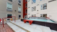 Located at 200 W Norwich Ave in Columbus, Ohio, this dynamic outdoor courtyard amenity space is nestled within a modern multi-story residential building. The courtyard is defined by a striking contemporary aesthetic, highlighted by bold red accents that carry throughout the furniture, railings, and architectural details, contrasting against the building's neutral beige and gray paneled exterior.

At the heart of the space is a sleek lap pool featuring an elegant waterfall wall clad in dark tile, accessible via stainless steel entry rails. Raised stone deck areas with step lighting surround the pool, providing elevated lounging zones with red geometric bench seating. Red powder-coated handrails guide transitions between levels, adding both safety and visual continuity.

Adjacent to the pool area is a lush artificial turf lawn, ideal for recreation, with a hammock stand and red-cushioned hammock as a central relaxation focal point. A cornhole board emblazoned with the Ohio state flag adds a local, community-oriented touch.

An expansive outdoor bar counter with a polished granite surface and red and white seating offers a social gathering hub, complemented by additional white lounge chairs. Planters with ornamental grasses and shrubs, young trees, and string lights overhead complete the inviting, resort-style atmosphere of this well-appointed communal courtyard.