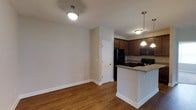 This single-story residence features three bedrooms and two bathrooms across a well-proportioned floor plan. The open-concept living and kitchen area serves as the heart of the home, where warm hardwood-style flooring extends throughout, complementing the neutral beige walls and bright white trim and baseboards.

The kitchen is outfitted with rich, dark wood cabinetry that provides ample storage, paired with granite countertops that add a refined touch. A peninsula with a breakfast bar separates the kitchen from the living area, offering additional prep space and casual seating. Black appliances, including a refrigerator, range, and over-the-range microwave, complete the kitchen's cohesive, contemporary look. Pendant lighting above the peninsula and ceiling-mounted fixtures provide warm, practical illumination throughout the space.

The adjacent living area is open and versatile, with a clean, uncluttered design that allows for flexible furniture arrangements. A paneled interior door leads to additional spaces within the home. The combination of durable flooring, neutral tones, and functional layout makes this residence a practical and comfortable living environment.