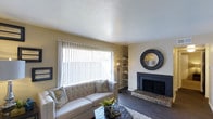 Located in Garland, this single-story home offers a thoughtfully designed layout that balances comfort and functionality. The living room serves as the heart of the space, showcasing dark hardwood floors that contrast beautifully with the warm, neutral-toned walls. A classic brick fireplace with a sleek black surround anchors one wall, providing a cozy focal point ideal for relaxed gatherings. A large picture window with vertical blinds floods the room with natural light, creating an open and airy atmosphere. Built-in shelving in the corner offers practical display space while adding architectural character to the room. The textured ceiling adds subtle dimension overhead, contributing to the room's inviting ambiance. The overall design aesthetic is clean and understated, blending traditional elements like the brick fireplace with contemporary touches, resulting in a space that feels both timeless and livable. The single-floor layout ensures seamless flow and accessibility throughout the home, making it a practical and welcoming residence in the Garland area.