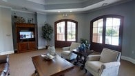 This single-floor space presents a refined and welcoming interior designed for comfort and elegance. The room is anchored by a cool blue-gray color palette on the walls, complemented by crisp white crown molding and a subtly recessed tray ceiling that adds architectural depth. Recessed lighting illuminates the space from above, enhancing the bright and airy atmosphere.

Two striking arched doorways with dark mahogany wood framing serve as prominent architectural focal points, each featuring multi-pane glass panels that allow natural light to pour in while offering views of an exterior courtyard or outdoor area. The arched design adds a classic, upscale character to the overall aesthetic.

The lounge area is furnished with cream-upholstered armchairs arranged around rich, dark wood coffee and accent tables, creating an inviting seating arrangement ideal for conversation or relaxation. Decorative accents, including teal ceramic vases and sculptural objects, are thoughtfully placed throughout. A large indoor tree adds a natural, organic element to the space, softening the formal décor.

A dark wood entertainment unit occupies one wall, providing both functional storage and visual grounding. The neutral carpeted flooring ties all elements together cohesively. Overall, the space exudes a polished, community-oriented ambiance suited to a clubhouse or common lounge setting.