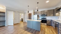 This well-appointed single-floor residence showcases a thoughtfully designed open-concept layout, where the kitchen serves as the centerpiece of the living space. The kitchen is outfitted with rich, gray-toned wood cabinetry, a full suite of stainless steel appliances including a refrigerator, range, microwave, and dishwasher, and a generous island topped with light granite countertops. Sleek black pendant lights hang above the island, adding a contemporary touch. A herringbone tile backsplash provides an elegant accent behind the sink area. Warm wood-look luxury vinyl plank flooring flows consistently throughout the main living areas, complementing the neutral gray wall tones and crisp white trim. The living area is bright and open, featuring a large sliding door with vertical blinds that invites natural light, alongside a built-in shelving nook for added functionality. The bathroom is finished with matching granite countertops, gray cabinetry, a framed mirror, and wood-look flooring, maintaining a cohesive aesthetic throughout. A spacious walk-in closet with wire shelving and soft carpet underfoot provides ample storage. Crown molding adds a refined detail to the living spaces, tying together the modern and classic elements of this carefully finished residence.